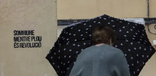 BURJASSOT (VALENCIA) COMUNIDAD VALENCIANA (REINO DE ESPAÑA, 29 DE SEPTIEMBRE DE 2025. Una mujer se protegía el lunes de la lluvia con un paraguas junto a una pintada con el lema "Sonreír mientras llueve es revolución", en Burjassot (Valencia). Efe