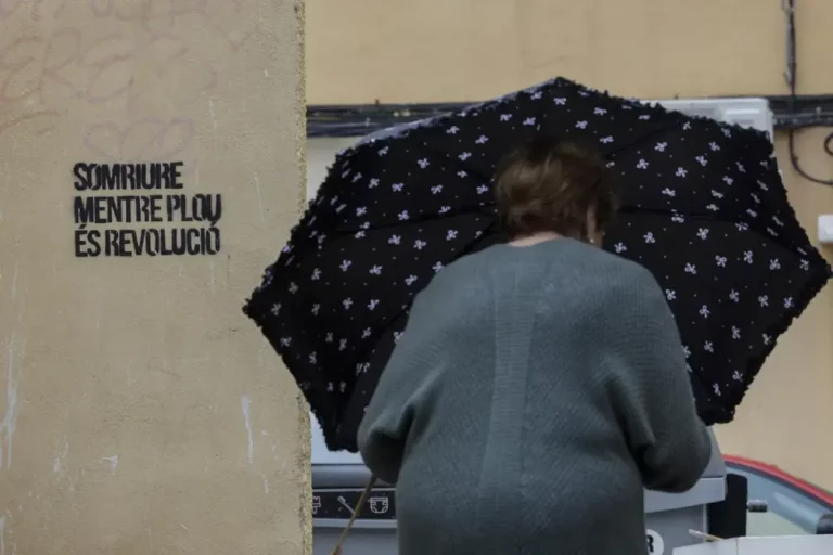 BURJASSOT (VALENCIA) COMUNIDAD VALENCIANA (REINO DE ESPAÑA, 29 DE SEPTIEMBRE DE 2025. Una mujer se protegía el lunes de la lluvia con un paraguas junto a una pintada con el lema "Sonreír mientras llueve es revolución", en Burjassot (Valencia). Efe