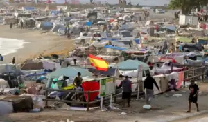 FOTOGRAFÍA. CEUTA, 26 DE SEPTIEMBRE DE 2026. Un inmenso y denso campamento clandestino levantado por miles de inmigrantes ilegales marroquíes abarrota por completo la arena, la zona rocosa y los accesos de la playa de El Trampolín de la Ciudad Autónoma de Ceuta, España. En el plano medio, sobresale una gran bandera de España colocada de forma llamativa sobre una de las tiendas de campaña de lona verde claro, junto a una valla de madera perimetral y carteles informativos del lugar. El asentamiento se extiende de forma masiva a lo largo de toda la línea de la costa, conformado por cientos de chabolas improvisadas construidas con plásticos de embalar, lonas multicolores, maderas, mantas y ropa tendida. En la parte inferior derecha, varios varones jóvenes transitan a pie por un camino de tierra adyacente a los habitáculos precarios, mientras el oleaje del mar rompe suavemente a la izquierda, evidenciando el desborde y la grave crisis de seguridad pública que se vive en este punto del litoral ceutí. EFE