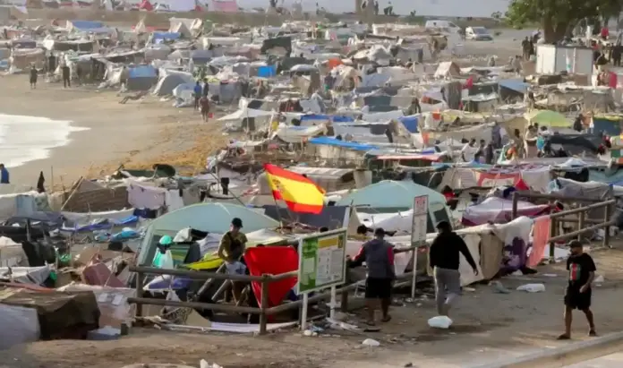 Campamento ilegal de inmigrantes, Detenidos dos inmigrantes marroquíes por violar a una menor en Ceuta FOTOGRAFÍA. CEUTA, 26 DE SEPTIEMBRE DE 2026. Un inmenso y denso campamento clandestino levantado por miles de inmigrantes ilegales marroquíes abarrota por completo la arena, la zona rocosa y los accesos de la playa de El Trampolín de la Ciudad Autónoma de Ceuta, España. En el plano medio, sobresale una gran bandera de España colocada de forma llamativa sobre una de las tiendas de campaña de lona verde claro, junto a una valla de madera perimetral y carteles informativos del lugar. El asentamiento se extiende de forma masiva a lo largo de toda la línea de la costa, conformado por cientos de chabolas improvisadas construidas con plásticos de embalar, lonas multicolores, maderas, mantas y ropa tendida. En la parte inferior derecha, varios varones jóvenes transitan a pie por un camino de tierra adyacente a los habitáculos precarios, mientras el oleaje del mar rompe suavemente a la izquierda, evidenciando el desborde y la grave crisis de seguridad pública que se vive en este punto del litoral ceutí. EFE