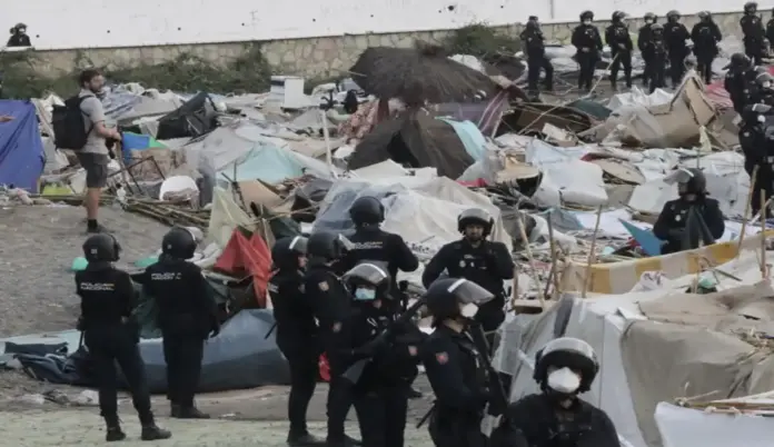 Desalojo masivo de inmigrantes en Ceuta por riesgo en una infraestructura estratégica FOTOGRAFÍA. CEUTA, 27 DE SEPTIEMBRE DE 2026. Un fuerte despliegue de agentes de la Unidad de Intervención Policial (UIP) de la Policía Nacional, equipados con cascos, protecciones y defensas reglamentarias, toma posiciones en pleno proceso de desmantelamiento del macrocampamento improvisado de inmigrantes ilegales de origen marroquí en la playa de Benítez de la Ciudad Autónoma de Ceuta, España. En el plano medio e inferior, se observa a un numeroso grupo de efectivos policiales coordinando el operativo a pie de campo; algunos de ellos portan mascarillas sanitarias y escopetas de dotación para el lanzamiento de material de seguridad. Detrás de la línea policial, se extiende una inmensa amalgama de chabolas, tiendas de campaña de plástico, lonas de colores azul y verde, mantas, cartones y sombrillas de playa destruidas que servían de refugio clandestino a los ocupantes. En la parte superior izquierda, un civil con mochila observa la intervención, mientras que al fondo, junto a un muro de piedra con pintadas, otra línea de agentes antidisturbios vigila el perímetro para asegurar el desalojo definitivo de esta zona adyacente a la planta desaladora. EFE