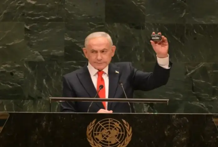 FOTOGRAFÍA. NUEVA YORK, 24 DE SÉPTIEMBRE DE 2026. El primer ministro del Estado de Israel, Benjamín "Bibi" Netanyahu, se dirige con ademán enérgico y semblante firme a la sesión plenaria de la 81ª Asamblea General de las Naciones Unidas. En la imagen, capturada desde la emblemática tribuna oficial del organismo internacional decorada en su parte frontal con el escudo de la ONU, el mandatario sostiene firmemente con su mano izquierda un pequeño dispositivo electrónico negro para ilustrar visualmente las amenazas tecnológicas y terroristas a las que se enfrenta la Tierra Santa, evidenciando su absoluto control de la escena frente al frustrado boicot de los sectores antisemitas. Crédito Lasvocesdelpueblo (Ñ Pueblo).
