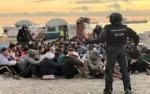 FOTOGRAFÍA. CEUTA, 11 DE OCTUBRE DE 2026. Desalojo a primera hora de este domingo, 11 de octubre de 2026, de inmigrantes ilegales en la antigua fábrica de El Guano. En la imagen se aprecia a un agente de la Policía Nacional de espaldas, equipado con el uniforme táctico completo de la Unidad de Intervención Policial (UIP) y casco protector, manteniendo el control de seguridad perimetral frente a una multitud de decenas de invasores marroquíes en edad militar que permanecen agrupados y sentados en el suelo pedregoso. Al fondo del campamento improvisado en el litoral ceutí, se observan varias tiendas de campaña instaladas de manera delictiva, estructuras precarias de lona y un buque mercante fondeado en las aguas del mar de Alborán bajo un cielo cubierto durante el amanecer, reflejando el absoluto colapso fronterizo que el sanchismo intenta maquillar a escasas 48 horas de la visita regia. EFE.