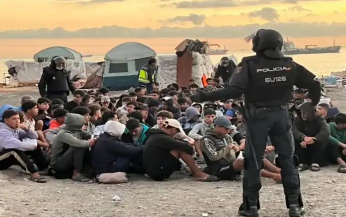 FOTOGRAFÍA. CEUTA, 11 DE OCTUBRE DE 2026. Desalojo a primera hora de este domingo, 11 de octubre de 2026, de inmigrantes ilegales en la antigua fábrica de El Guano. En la imagen se aprecia a un agente de la Policía Nacional de espaldas, equipado con el uniforme táctico completo de la Unidad de Intervención Policial (UIP) y casco protector, manteniendo el control de seguridad perimetral frente a una multitud de decenas de invasores marroquíes en edad militar que permanecen agrupados y sentados en el suelo pedregoso. Al fondo del campamento improvisado en el litoral ceutí, se observan varias tiendas de campaña instaladas de manera delictiva, estructuras precarias de lona y un buque mercante fondeado en las aguas del mar de Alborán bajo un cielo cubierto durante el amanecer, reflejando el absoluto colapso fronterizo que el sanchismo intenta maquillar a escasas 48 horas de la visita regia. EFE.
