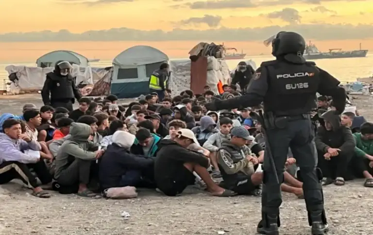 FOTOGRAFÍA. CEUTA, 11 DE OCTUBRE DE 2026. Desalojo a primera hora de este domingo, 11 de octubre de 2026, de inmigrantes ilegales en la antigua fábrica de El Guano. En la imagen se aprecia a un agente de la Policía Nacional de espaldas, equipado con el uniforme táctico completo de la Unidad de Intervención Policial (UIP) y casco protector, manteniendo el control de seguridad perimetral frente a una multitud de decenas de invasores marroquíes en edad militar que permanecen agrupados y sentados en el suelo pedregoso. Al fondo del campamento improvisado en el litoral ceutí, se observan varias tiendas de campaña instaladas de manera delictiva, estructuras precarias de lona y un buque mercante fondeado en las aguas del mar de Alborán bajo un cielo cubierto durante el amanecer, reflejando el absoluto colapso fronterizo que el sanchismo intenta maquillar a escasas 48 horas de la visita regia. EFE.
