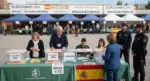 FOTOGRAFÍA. MADRID, 29 NOVIEMBRE 2024. Vista en primer plano de una mesa electoral dispuesta en el patio exterior de un polideportivo municipal durante la jornada de votación de las Elecciones 29N. En la escena principal, se observa con total nitidez a los miembros de la Junta Electoral Central debidamente sentados y gestionando las actas, mientras varios ciudadanos introducen ordenadamente sus sobres en urnas de metacrilato transparente serigrafiadas con la inscripción "Elecciones 29N". El recinto se encuentra fuertemente custodiado por agentes uniformados de las Fuerzas y Cuerpos de Seguridad del Estado (Policía Nacional y Guardia Civil), quienes garantizan el correcto desarrollo del proceso democrático en un ambiente pacífico y seguro. Al fondo, bajo carpas y pancartas oficiales, una gran muchedumbre de votantes aguarda en largas filas su turno para ejercer el derecho al voto. IA/Lasvocesdelpueblo (Ñ Pueblo).