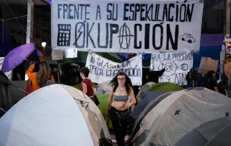 FOTOGRAFÍA, MADRID, 05 DE OCTUBRE DE 2026. Una abigarrada multitud de militantes y activistas de ultraizquierda abarrota la totalidad de la remodelada Puerta del Sol de Madrid, concentrándose de manera masiva en las inmediaciones de la Real Casa de Correos. Los manifestantes sostienen en alto una gran pancarta central de lona blanca donde se lee en letras negras y rojas el lema "Derecho a techo: Viva la Okupación y huelga de alquileres contra los rentistas", flanqueada por numerosas banderas de color rojo, símbolos comunistas y enseñas republicanas que ondean sobre las cabezas de la multitud. En el plano medio, se aprecian decenas de tiendas de campaña de estilo "iglú" en tonos verdes, azules y grises, instaladas directamente sobre las losas de granito de la plaza, rodeando las salidas acristaladas de la estación de Cercanías del Sol y la estatua de Carlos III. Rostros de jóvenes y activistas veteranos con megáfonos rompen la línea del fondo, donde varias pancartas secundarias exigen la "Inquiokupación legítima frente a los desahucios" y la "expropiación de fondos buitre", conformando una estampa de ocupación ilegal del espacio público custodiada a la distancia por varias furgonetas azules de las Unidades de Intervención Policial (UIP) de la Policía Nacional. EFE