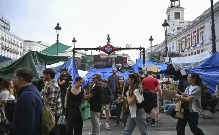 FOTOGRAFÍA. MADRID, 16 DE OCTUBRE DE 2026. Vista de la entrada principal de la estación de Metro de Sol capturada durante las horas previas a la clausura total decretada por la Comunidad de Madrid a causa del grave problema de insalubridad y acumulación de orines provocado por la extrema izquierda. La imagen documenta la degradación total del kilómetro cero de la capital, mostrando el acceso al suburbano completamente sitiado y rodeado por decenas de lonas de plástico azul y verde, estructuras precarias y tiendas de campaña pertenecientes a la acampada ilegal que okupa la plaza desde hace tres semanas. Varios agentes de la Policía Nacional observan la escena en un lateral, mientras un flujo constante de pasajeros, trabajadores y ciudadanos de a pie se ven obligados a sortear los residuos orgánicos, carteles ideológicos y barricadas de los manifestantes antisistema para poder acceder a los andenes, en un ambiente que los informes de riesgos laborales declararon impracticable por los altos niveles de amoníaco detectados en los pozos de ventilación exteriores. EFE