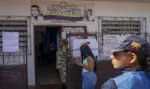 FOTOGRAFÍA. CARACAS, 18 DE OCTUBRE DE 2026. Una pancarta con el lema «Cerritos Unidos Chávez por Siempre» y los rostros de Hugo Chávez y Nicolás Maduro corona la entrada de la casa comunal Doctor Duque, habilitada como centro de votación con el cartel del CNE, mientras en el exterior una agente uniformada con traje militar de camuflaje permanece de pie en la puerta, una joven con camiseta azul claro y vaqueros consulta su teléfono móvil y, en primer plano a la derecha, una funcionaria con gorra con la bandera de Venezuela y chaleco azul observa el desarrollo de la jornada. EFE
