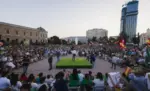 FOTOGRAFÍA. MADRID, 21 DE JULIO DE 2023. El presidente de Vox, Santiago Abascal Conde, interviene de espaldas al objetivo sobre una estructura o tarima cuadrada de color verde situada en el centro de la emblemática Plaza de Colón de Madrid, protagonizando el multitudinario mitin de cierre de campaña de las elecciones generales de 2023. El líder patriota se encuentra arropado en el escenario por destacados miembros de la cúpula nacional y regional del partido, mientras una marea humana compuesta por miles de simpatizantes y militantes abarrota por completo la explanada, rodeando la zona central tanto sentados en hileras de sillas como de pie. En la masiva concentración ondean numerosos estandartes, destacando una gran profusión de banderas de España y enseñas verdes de Vox que colorean el ambiente. En el primer plano, parte del público asistente inmortaliza el momento con sus teléfonos móviles o sostiene pancartas y banderas, mientras que al fondo se divisan edificios históricos de la capital junto a modernas infraestructuras urbanas y un gran rascacielos en construcción bajo un cielo despejado de atardecer. Lasvocesdelpueblo (Ñ Pueblo)