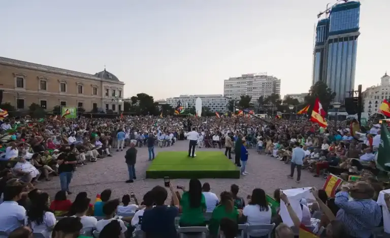 FOTOGRAFÍA. MADRID, 21 DE JULIO DE 2023. El presidente de Vox, Santiago Abascal Conde, interviene de espaldas al objetivo sobre una estructura o tarima cuadrada de color verde situada en el centro de la emblemática Plaza de Colón de Madrid, protagonizando el multitudinario mitin de cierre de campaña de las elecciones generales de 2023. El líder patriota se encuentra arropado en el escenario por destacados miembros de la cúpula nacional y regional del partido, mientras una marea humana compuesta por miles de simpatizantes y militantes abarrota por completo la explanada, rodeando la zona central tanto sentados en hileras de sillas como de pie. En la masiva concentración ondean numerosos estandartes, destacando una gran profusión de banderas de España y enseñas verdes de Vox que colorean el ambiente. En el primer plano, parte del público asistente inmortaliza el momento con sus teléfonos móviles o sostiene pancartas y banderas, mientras que al fondo se divisan edificios históricos de la capital junto a modernas infraestructuras urbanas y un gran rascacielos en construcción bajo un cielo despejado de atardecer. Lasvocesdelpueblo (Ñ Pueblo)
