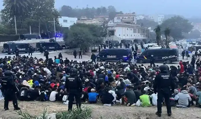 FOTOGRAFÍA. CEUTA, 04 DE OCTUBRE DE 2026. Centenares de inmigrantes ilegales marroquíes permanecen agrupados y custodiados en el suelo por los efectivos de la Policía Nacional durante el despliegue del macrooperativo policial y militar activado a primeras horas de este domingo para desmantelar el asentamiento ilegal de la playa de El Trampolín de Ceuta. En la imagen se aprecia la magnitud del dispositivo de seguridad, con agentes de las Unidades de Intervención Policial (UIP) rodeando a los indocumentados y una hilera de furgones policiales blindados bloqueando los viales y accesos a la zona costera, mientras al fondo de la playa se observan los campamentos de lonas y estructuras precarias levantados durante los 66 días de invasión masiva. EFE