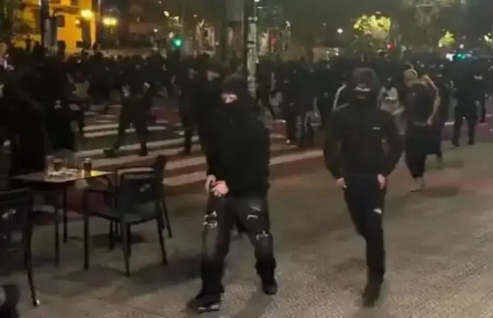La infantería del islamoizquierdismo y filoterrorismo etarra queman Bilbao (1) FOTOGRAFÍA. DEUSTO (BILBAO), 26 DE OCTUBRE DE 2026. La imagen muestra a un grupo nutrido de delincuentes y violentos parapetados en la oscuridad de la noche en pleno casco urbano de Bilbao, bloqueando el tránsito en un paso de peatones. En primer término destacan dos agitadores fuertemente encapuchados que avanzan desafiantes hacia la cámara. Sujeto de la izquierda: Lleva el rostro completamente cubierto por un pasamontañas negro que solo deja al descubierto la zona de los ojos. Viste una sudadera deportiva negra de manga larga con capucha ajustada sobre la cabeza, pantalones oscuros de chándal con estampados/letras blancas de estilo urbano y calzado deportivo oscuro. Camina en postura amenazante, manteniendo las manos en la parte frontal. Sujeto de la derecha: Avanza de forma coordinada luciendo un atuendo idéntico de guerrilla urbana: sudadera negra de capucha sobre la cabeza, pasamontañas/mascarilla negra que oculta totalmente sus facciones, pantalones de chándal oscuros ajustados y zapatillas deportivas negras. Entorno y fondo: Al fondo se observa una muchedumbre de decenas de extremistas enfundados en ropajes negros y capuchas invadiendo la calzada y las aceras. A la izquierda, en primer plano, se distinguen veladores y sillas de la terraza de un establecimiento hostelero desierta tras el asedio, así como la señalización luminosa de los semáforos en verde e iluminación urbana. Lasvocesdelpueblo (Ñ Pueblo)