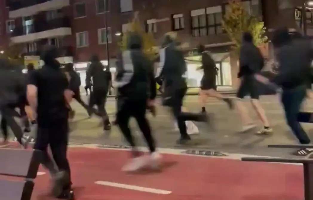 FOTOGRAFÍA. DEUSTO (BILBAO), 26 DE OCTUBRE DE 2026. Descripción minuciosa: La imagen capta el momento exacto en que una turba de radicales enmascarados emprende la huida a la carrera tras sembrar el caos en la vía pública, cruzando un carril bici rojo delimitado en la calzada. Indumentaria y perfiles: Todos los individuos que integran el grupo visten ropa de camuflaje urbano y deporte en tonos oscuros (negro, gris y azul marino) para dificultar su identificación. La media de edad aparente se sitúa entre los 18 y los 25 años. Sujeto en primer plano a la izquierda: Se aprecia a un individuo de espaldas vistiendo camiseta negra de manga corta, pantalones oscuros ajustados y calzado deportivo negro, corriendo activamente hacia el frente. Sujetos centrales en carrera: Varios delincuentes huyen a gran velocidad portando sudaderas con capucha (hoodies) negras y grises —algunas con franjas blancas en las mangas de estilo Adidas—, pantalones de chándal y bermudas oscuras sobre mallas deportivas. En la parte posterior se observa a uno de ellos corriendo con pantalón corto y zapatillas de deporte con calcetines claros. Fondo y escenario de los altercados: En la parte posterior se aprecia la fachada de edificios residenciales de varias alturas con luces encendidas en las ventanas, comercios a pie de calle e iluminación pública, evidenciando que la persecución y los disturbios tuvieron lugar en plenas zonas residenciales y comerciales del centro urbano. Lasvocesdelpueblo (Ñ pueblo)