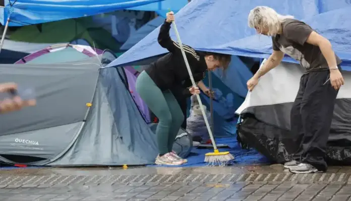 FOTOGRAFÍA. MADRID, 06 DE OCTUBRE DE 2026. Dos jóvenes integrantes de la acampada ilegal ultraizquierdista instalada en la Puerta del Sol retiran el agua acumulada sobre las lonas y tiendas de campaña tras el intenso temporal de lluvia y fuertes rachas de viento registrado este martes en la capital. En la imagen se aprecia en primer plano a una activista con mallas verdes barriendo el agua con una escoba sobre una lona azul, mientras su compañero, con ropa oscura, sujeta los techos de una de las estructuras provisionales para evitar daños mayores en el campamento. Los colectivos extremistas Sindicato de Inquilinas y Sindicato de Estudiantes, que defienden la impunidad de la okupación y la inquiokupación, se han visto obligados a alterar las asambleas previstas para la jornada debido a las inundaciones en sus puestos, aunque mantienen sus planes de resistencia frente a un posible desalojo policial y continúan con la organización de una inminente huelga general. EFE