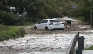 FOTOGRAFÍA. RONDA SUR DE GRANOLLERS, 05 DE OCTUBRE DE 2026. Un coche de color blanco de la marca Peugeot ha quedado completamente atrapado en mitad de una densa riada de barro y lodo que ha anegado la calzada tras las intensas precipitaciones acumuladas en las últimas horas en Cataluña. Junto a la parte trasera del turismo, un vecino con pantalones cortos intenta retirar desesperadamente los sedimentos acumulados con la ayuda de una pala, tratando de liberar las ruedas del vehículo en un entorno rural rodeado de cañas y vegetación salvaje donde se aprecia una señal de tráfico de carril bici. El terreno muestra los estragos de las inundaciones provocadas por el temporal que mantiene activado el plan Inuncat, con el camino totalmente cubierto por una capa de lodo marrón y un cordón policial en primer término que prohíbe el paso a la zona afectada. EFE