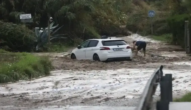 FOTOGRAFÍA. RONDA SUR DE GRANOLLERS, 05 DE OCTUBRE DE 2026. Un coche de color blanco de la marca Peugeot ha quedado completamente atrapado en mitad de una densa riada de barro y lodo que ha anegado la calzada tras las intensas precipitaciones acumuladas en las últimas horas en Cataluña. Junto a la parte trasera del turismo, un vecino con pantalones cortos intenta retirar desesperadamente los sedimentos acumulados con la ayuda de una pala, tratando de liberar las ruedas del vehículo en un entorno rural rodeado de cañas y vegetación salvaje donde se aprecia una señal de tráfico de carril bici. El terreno muestra los estragos de las inundaciones provocadas por el temporal que mantiene activado el plan Inuncat, con el camino totalmente cubierto por una capa de lodo marrón y un cordón policial en primer término que prohíbe el paso a la zona afectada. EFE