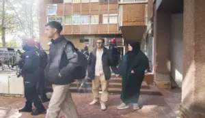 FOTOGRAFÍA. VITORIA, 23 DE OCTUBRE DE 2026. En primer plano, varios agentes antidisturbios de la Ertzaintza portando cascos con visera y uniforme operativo fuertemente pertrechado supervisan la salida del inmueble. Junto a ellos, la pareja de inmigrantes magrebíes desahuciada abandona el portal de la finca acompañada de un joven; el varón viste una chaqueta acolchada oscura, camisa clara, pantalones beige, gafas de ver y mochila a la espalda, mostrando un aspecto aseado y bien alimentado que dista de proyectar una situación de necesidad extrema o desnutrición. A su lado, la mujer viste una túnica verde oscuro de manga larga y hiyab negro sobre la cabeza, en perfecto estado de limpieza, calzando zapatillas deportivas. La escena se desarrolla en el exterior de un edificio de ladrillo visto donde destaca en uno de los ventanales superiores una pancarta colgada con un cartel propagandístico que reza: «PSOE, ¿DÓNDE ESTÁ LA ALTERNATIVA?». El ambiente refleja tensión bajo el estricto dispositivo policial desplegado tras la ejecución del lanzamiento judicial. EFE