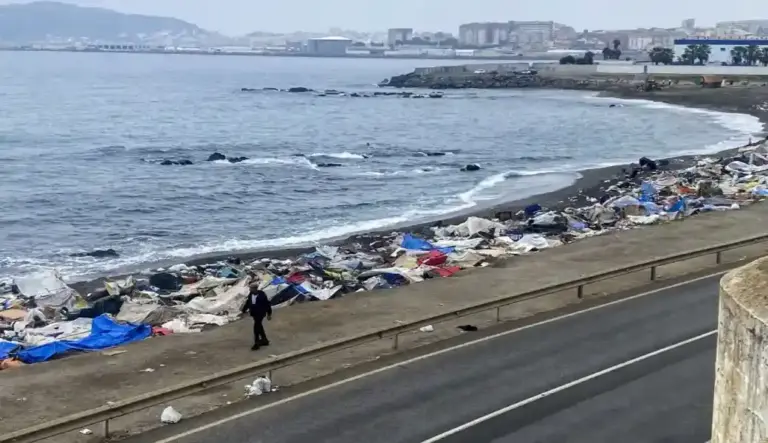 FOTOGRAFÍA. CEUTA, 06 DE OCTUBRE DE 2026. Una deprimente hilera de infraviviendas provisionales, tiendas de campaña rudimentarias y chabolas construidas de forma ilegal con lonas azules, plásticos blancos y mantas se extiende a lo largo de toda la orilla de arena negra de la Playa de El Trampolín en Ceuta, evidenciando el rastro de suciedad y degradación tras el asalto masivo perpetrado a pie y a nado por miles de inmigrantes ilegales marroquíes a finales de julio. En el plano medio, un hombre solitario vestido con ropa oscura camina junto a los restos del asentamiento desmantelado, delimitado en primer término por una carretera y un quitamiedos metálico donde se acumulan bolsas de basura y desperdicios. Al fondo, las aguas de la bahía norte se baten contra las rocas bajo un cielo completamente cubierto y gris, recortándose en el horizonte la silueta urbana de la ciudad autónoma y el puerto, donde maquinaria pesada trabaja en la limpieza de la línea de costa custodiada por las Fuerzas y Cuerpos de Seguridad del Estado. EFE
