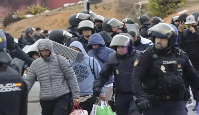 Policía y Guardia Civil van a por las casas-patera que ocultan invasores en Ceuta FOTOGRAFÍA. CEUTA, 18 DE OCTUBRE DE 2026. Varios agentes de la Unidad de Intervención Policial (UIP) de la Policía Nacional, provistos de cascos, chalecos antibalas y equipo antidisturbios, custodian y escoltan a una columna de inmigrantes ilegales jóvenes que portan sacos de dormir, esterillas, mochilas y bolsas con sus enseres personales durante el operativo de desalojo de los asentamientos y naves del Tarajal, en Ceuta. EFE