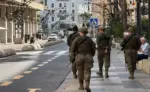 FOTOGRAFÍA. CEUTA, 18 DE OCTUBRE DE 2026. Cuatro militares del Ejército de Tierra ataviados con uniforme de campaña, boina y armamento a la espalda patrullan a pie por una céntrica acera de la ciudad autónoma de Ceuta, bajo la mirada de un ciudadano mayor sentado en un banco de piedra, en una calle custodiada por banderas de España y edificios con soportales. EFE
