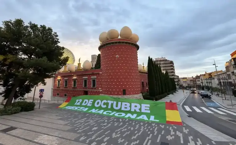 FOTOGRAFÍA. FIGUERAS, 12 OCTUBRE DE 2026. La instantánea retrata los exteriores de la plaza que da acceso a la Torre Galatea del Teatro-Museo Dalí de Figueras, donde militantes y simpatizantes de la formación patriota Vox han desplegado una imponente lona de color verde de varios metros de longitud que reza en letras blancas capitulares «12 DE OCTUBRE. FIESTA NACIONAL», flanqueada en sus dos extremos por los colores de la enseña nacional de España. Al fondo se recorta el singular edificio de color granate coronado por sus emblemáticos huevos gigantes y la cúpula geodésica acristalada diseñada por el genio surrealista Salvador Dalí, contrastando con la calzada limpia y semivacía de la avenida adyacente durante el amanecer de este lunes. La acción de guerrilla pacífica enmarcada en la declaración institucional del diputado Alberto Tarradas Peneque escenifica la resistencia cultural de la formación verde en el corazón del independentismo de cara a las próximas elecciones del 29 de noviembre, proyectando un mensaje nítido de soberanía, orgullo e indisoluble unidad nacional. Lasvocesdelpueblo (Ñ Pueblo).