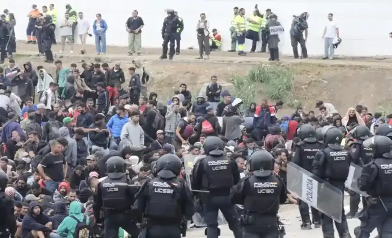 FOTOGRAFÍA. CEUTA, 04 DE OCTUBRE DE 2026. Centenares de inmigrantes ilegales de la invasión marroquí permanecen agrupados y fuertemente custodiados por efectivos de las Unidades de Intervención Policial (UIP) de la Policía Nacional durante el macrooperativo desplegado este domingo para el desalojo forzoso del asentamiento ilegal de la playa de El Trampolín de Ceuta. En la imagen se aprecia un numeroso grupo de indocumentados vigilados en primer término por una barrera de agentes con cascos, defensas y escudos protectores transparentes, mientras al fondo, en una zona elevada junto a un muro blanco, civiles, personal de emergencias y más dotaciones policiales controlan el perímetro de seguridad para evitar fugas. EFE